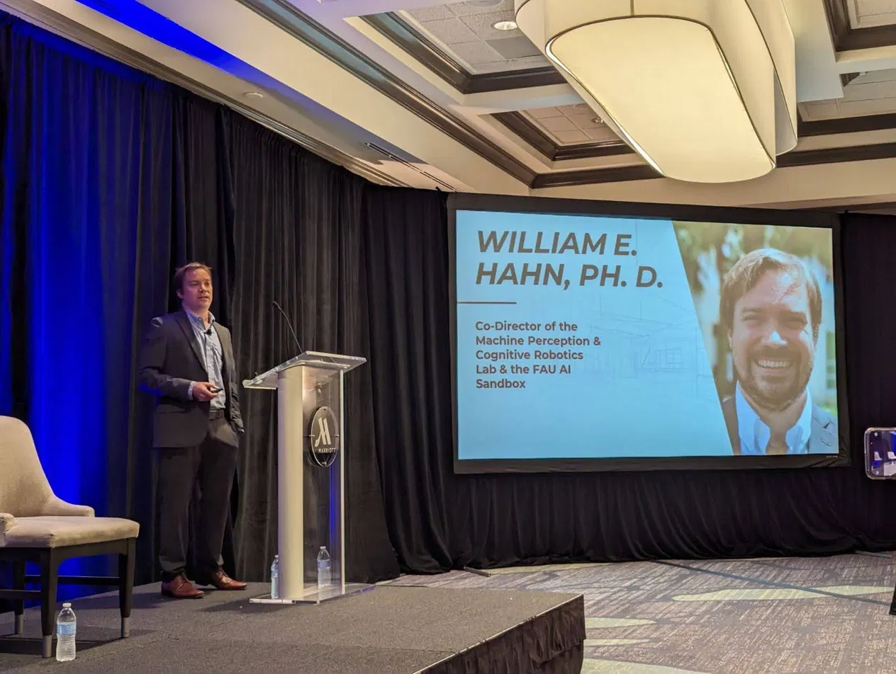 Dr. William Hahn on stage beside a screen showing his title: Co-Director of the Machine Perception and Cognitive Robotics Lab and the FAU AI Sandbox